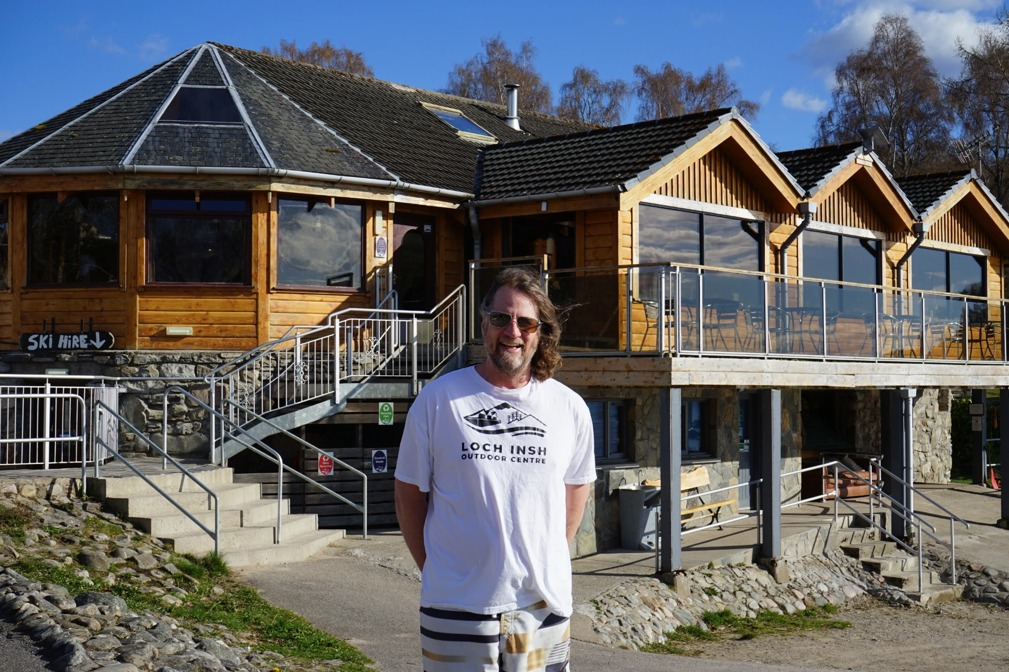 Man in glasses and a white "Loch Insh Outdoor Centre" shirt smiles outside a wooden building with large windows and stairs. The sky is clear and sunny.
