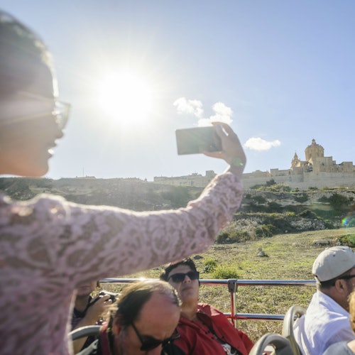 Человек в розовой кружевной блузке делает селфи со смартфоном; на заднем плане живописный вид с зданием.