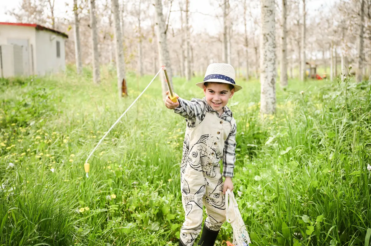 צילום קונספט יצירתי של ילד קטן ומתוק לבוש כדייג עם חכה קטנה וכובע קש, מחייך בלב חורשה ירוקה ופורחת. התמונה משדרת תמימות ושמחה של ילדות בטבע, מאת Shiriz Photography והצלמת שירי עציץ