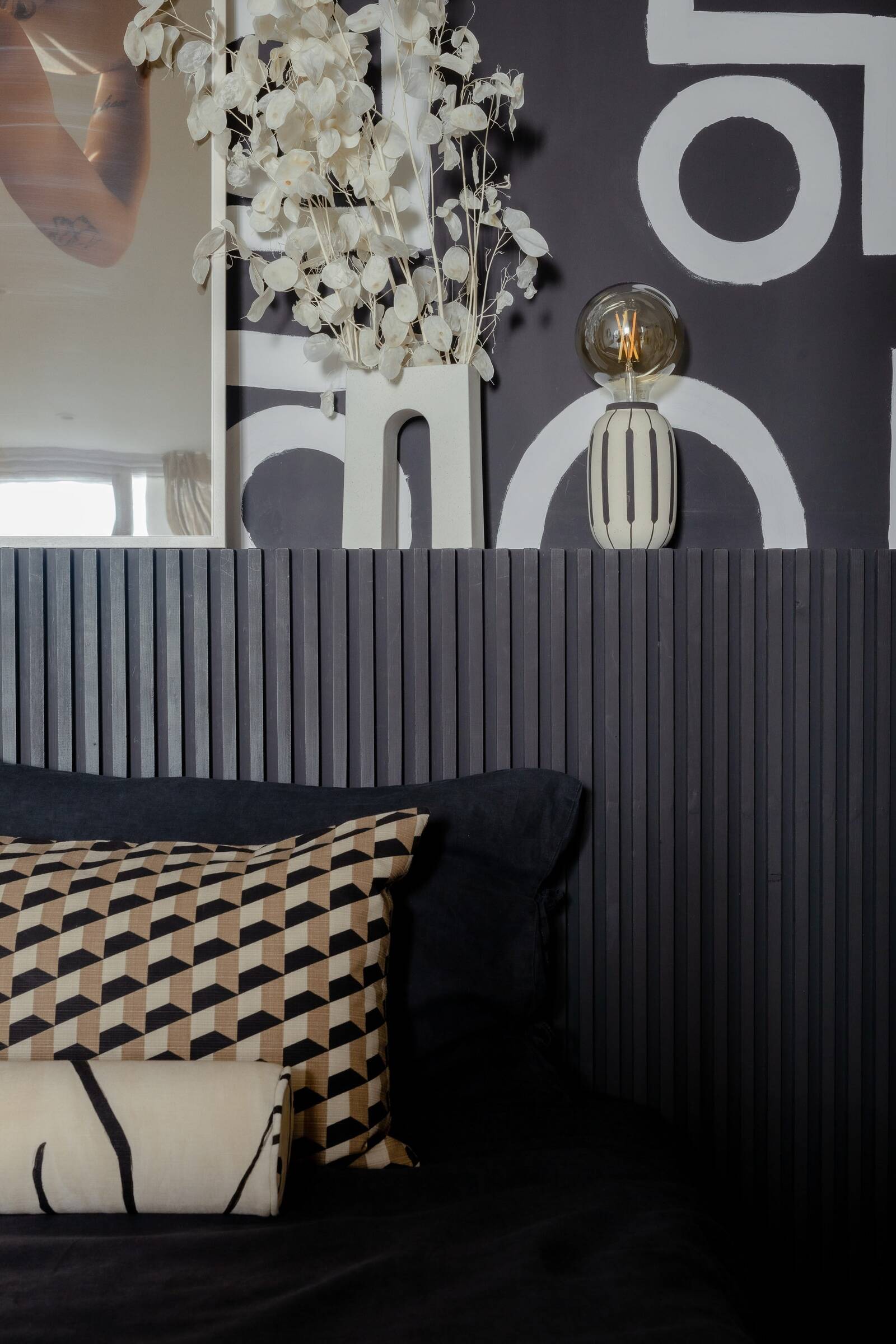 Interior view of a bed with patterned pillows against a dark, textured headboard. Behind the bed, there's a decorative shelf with a glass-encased light and a ceramic vase with dried white foliage, a mirror, and a partial view of a window with curtains.