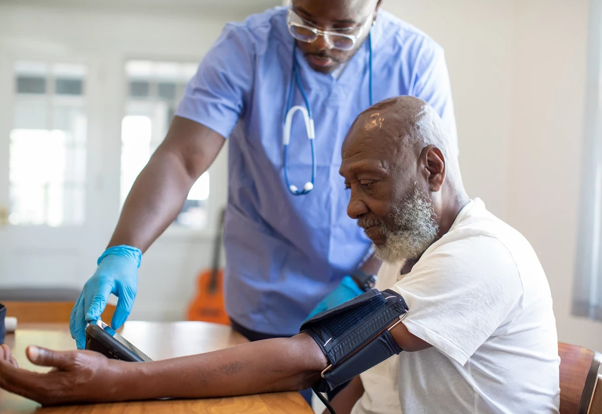 Healthcare professional wearing gloves and scrubs measures a patient’s blood pressure using an arm cuff and monitor.