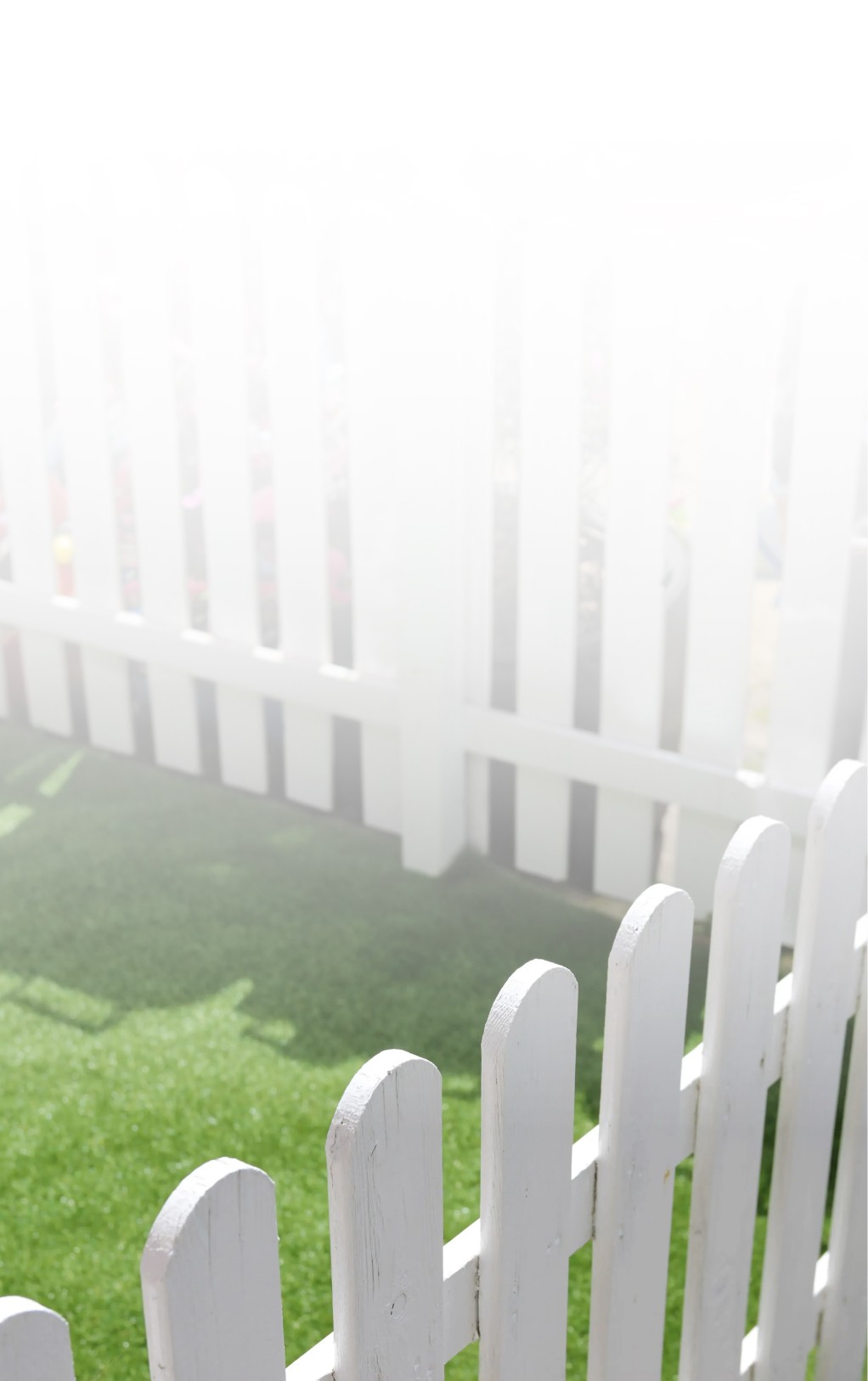 A sunlit, white picket fence casts gentle shadows on a vibrant green lawn, creating a serene and inviting outdoor setting.