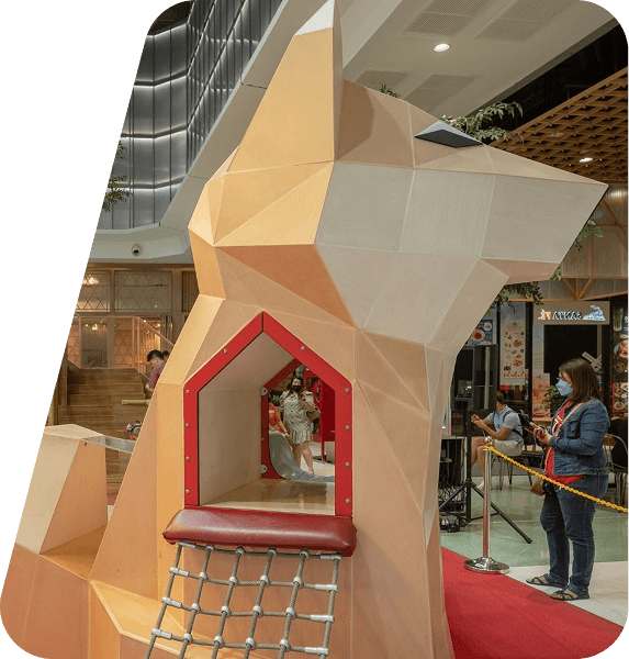 Colorful playground with intricate climbing structures, including a netted white and orange dome, set on a bright, winding path. Lush greenery surrounds the area, giving it an inviting and vibrant atmosphere.
