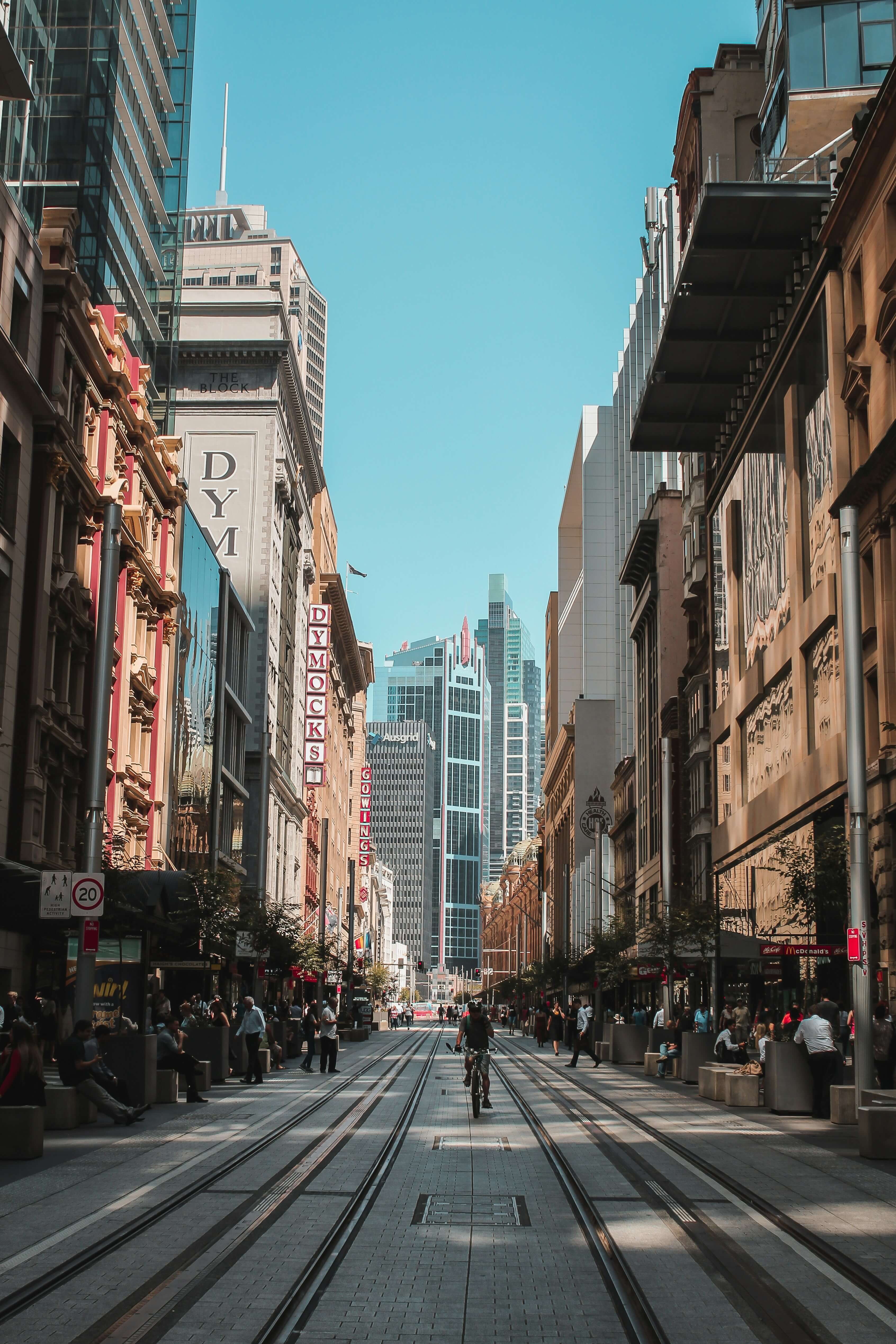 A cyclist rides down a busy city street lined with tall buildings, shops, and pedestrians under a clear blue sky.