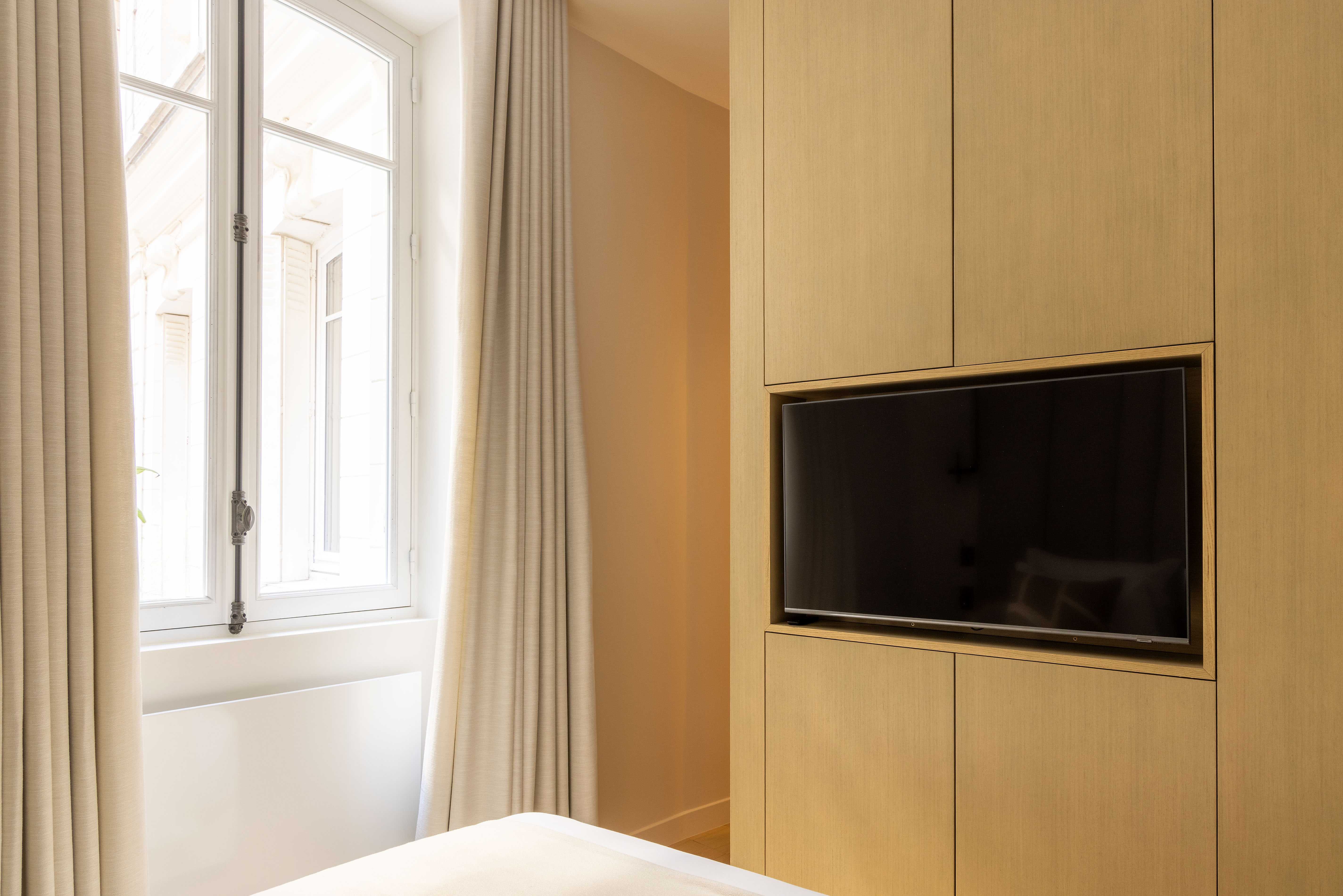 Chambre avec télévision encastrée dans meuble en bois clair, fenêtre et rideaux beiges