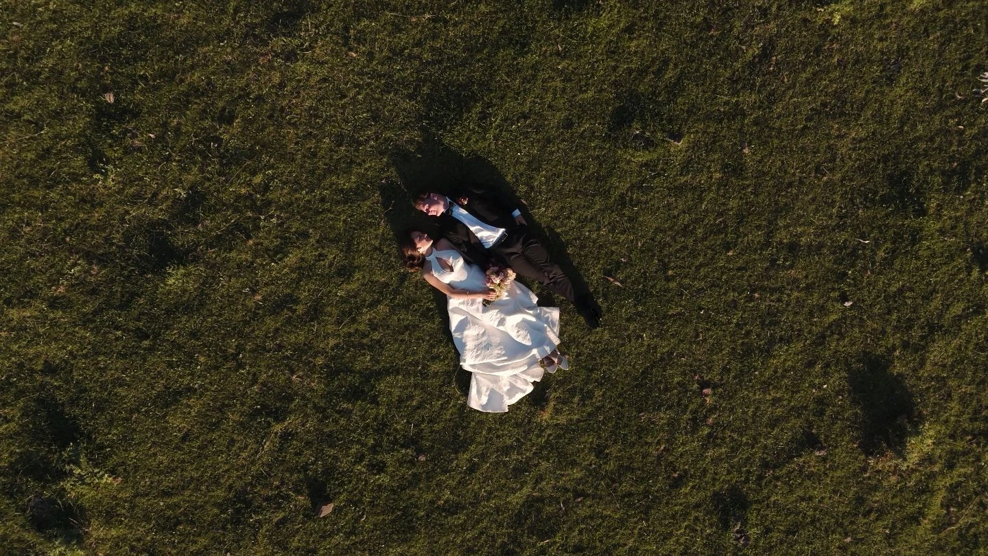 Aerial view of a bride and groom lying on a grassy field, with the bride in a white gown holding a bouquet and the groom in a dark suit, surrounded by lush green grass under natural sunlight.