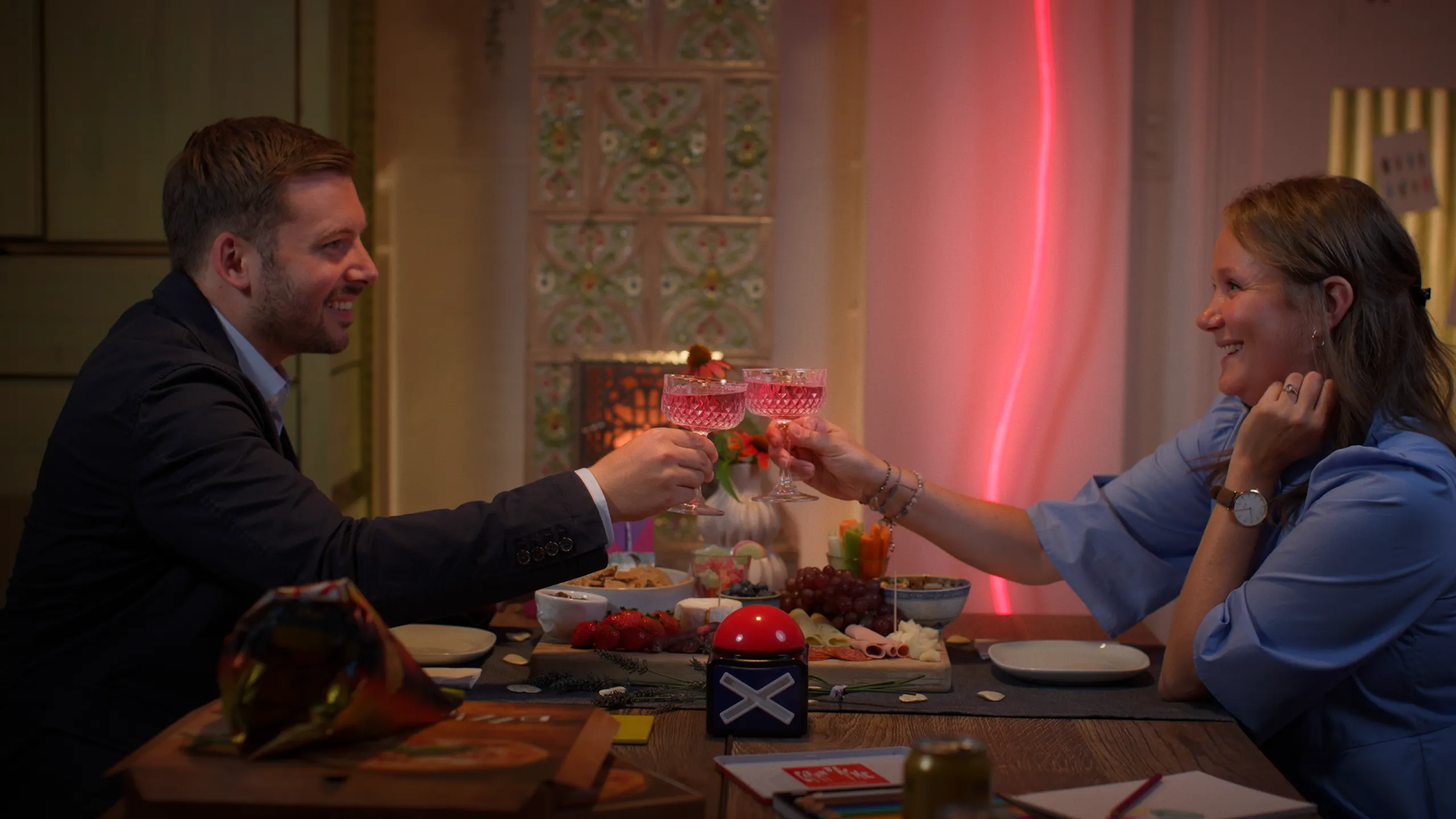 A smiling man and woman at a dinner table toasting with pink cocktails in a warmly lit room.