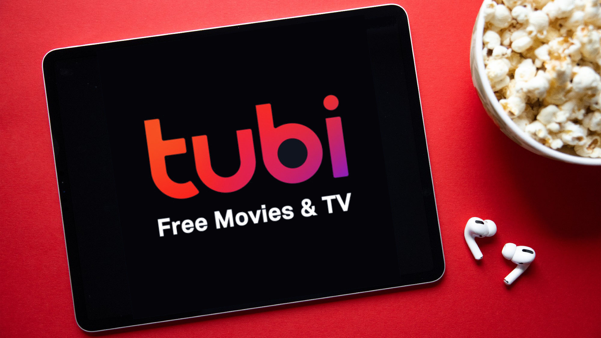 The word "Tubi" is displayed boldly, with "Free Movies and TV" below it, all on a black square against a red background. In the top right corner of the red background, there’s a bowl of popcorn, and at the bottom, two AirPods are visible. 