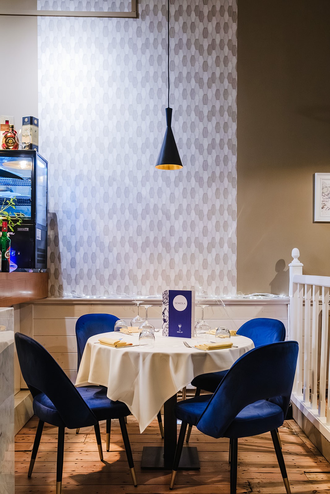 A neatly set round dining table with a cream tablecloth, surrounded by deep blue velvet chairs. A pendant light hangs above, and the table is arranged with folded napkins, upside-down glasses, cutlery and a Semola wine list placed at the centre, against a patterned grey wall with soft decorative lighting.