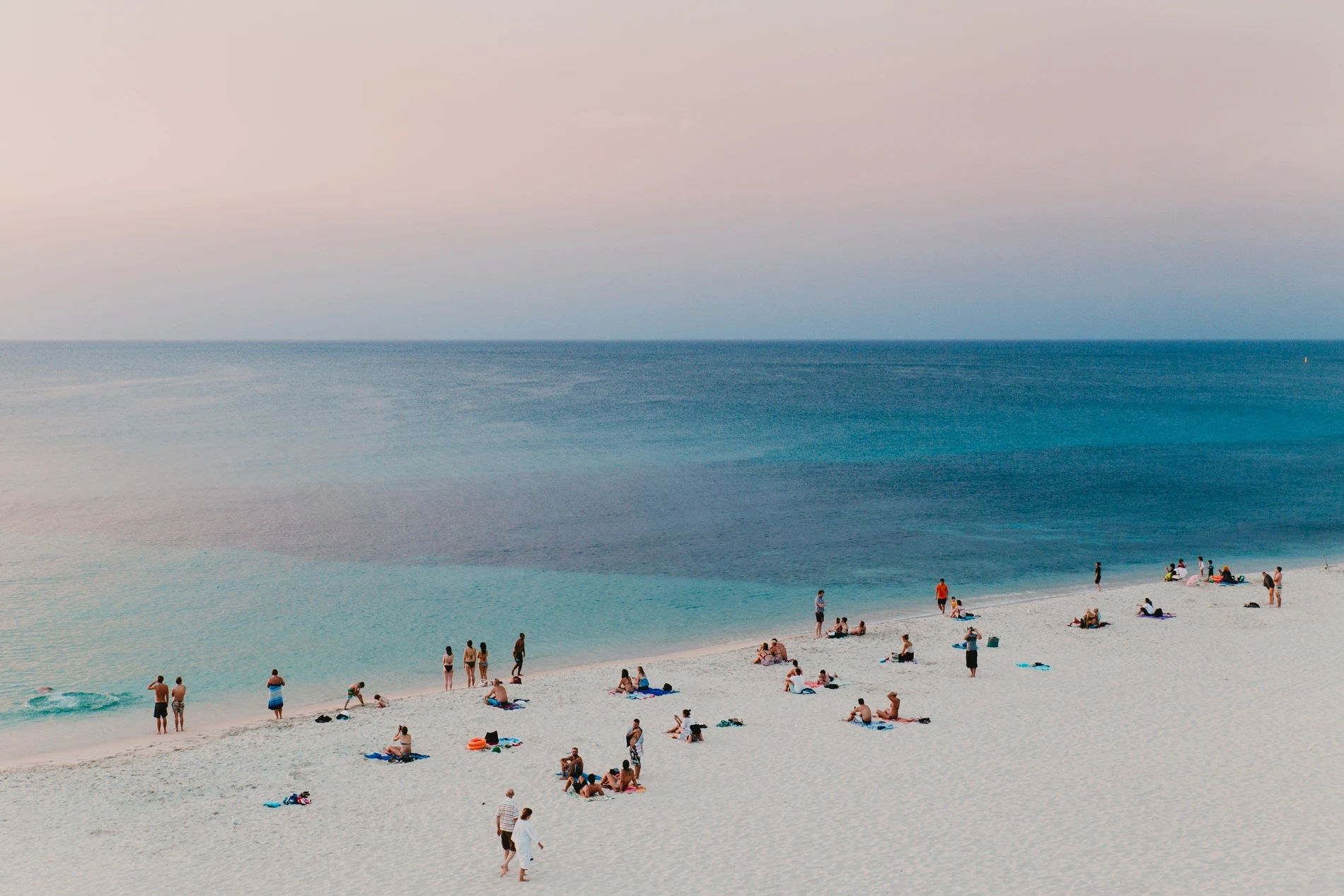 A busy white sand beach seen from above in soft pastel light, swimmers scattered through the shallows