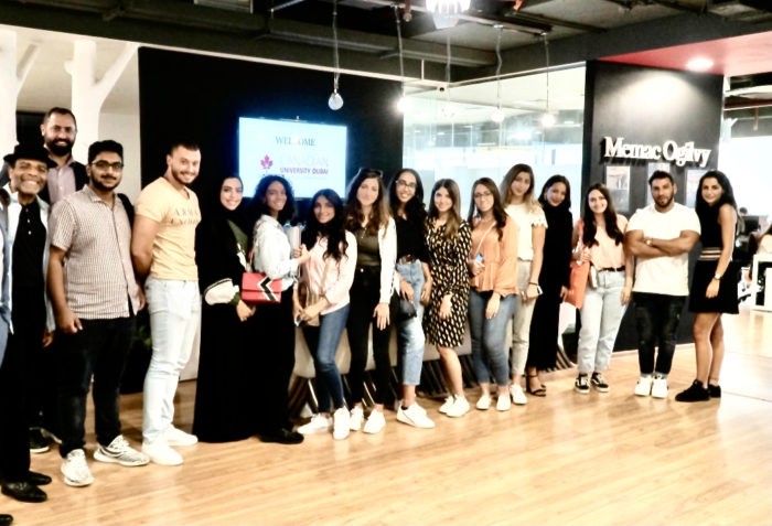 A group of young professionals stands in a line inside a modern office with wood flooring and glass walls; a screen displays "Metaquery" and "British Bank" logos in the background.