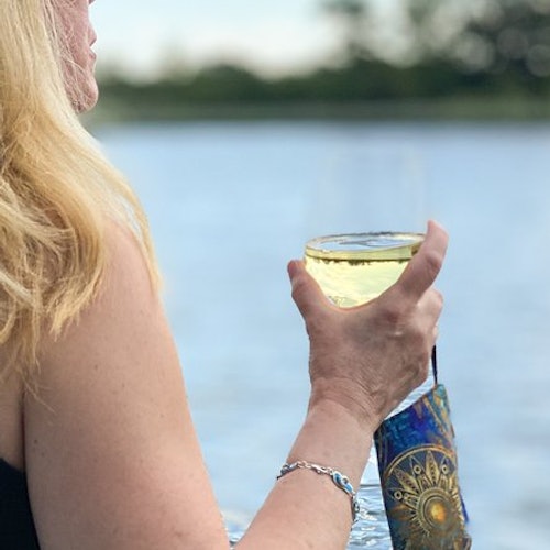 Person with long blonde hair holding a yellow drink and a colorful face mask, standing near a body of water. Trees are in the background.