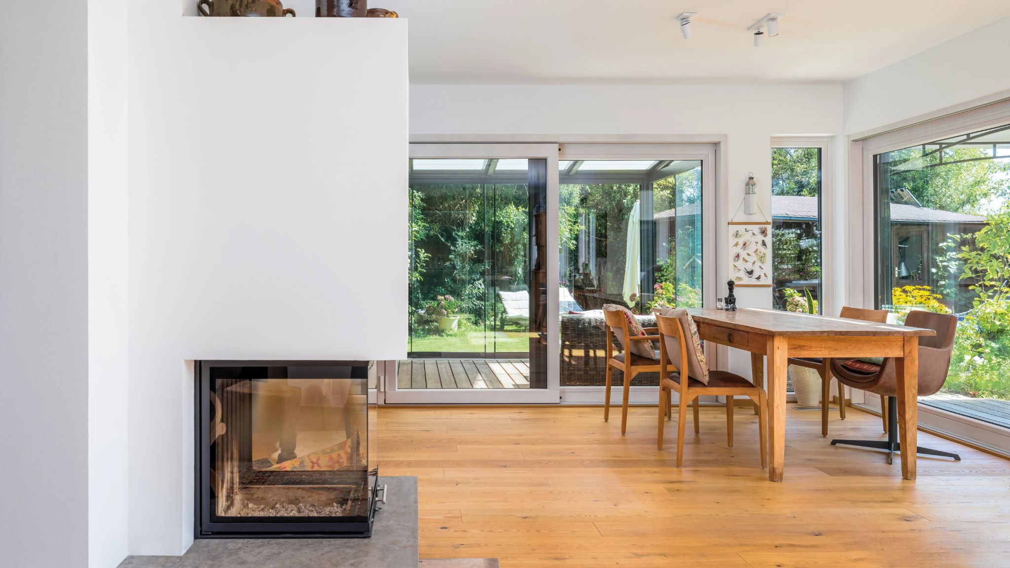 Moderner Wohnbereich mit Kamin, Holzboden und Essbereich, der auf einen überdachten Garten mit Terrassentüren blickt.