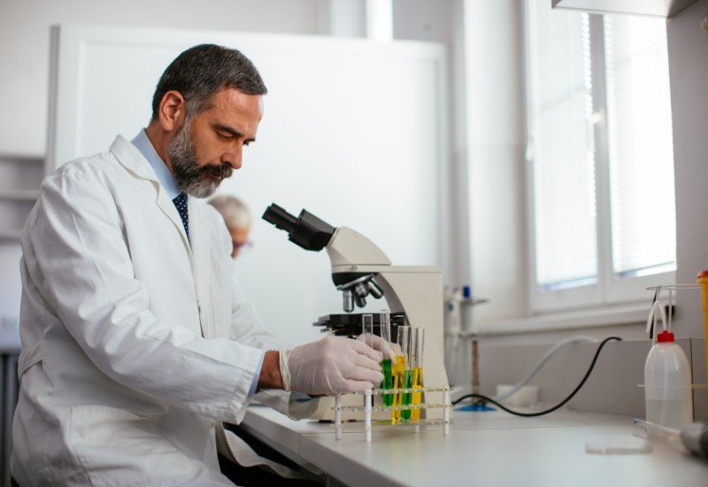 Male clinician in a laboratory examining test tubes beside a microscope during pathology testing