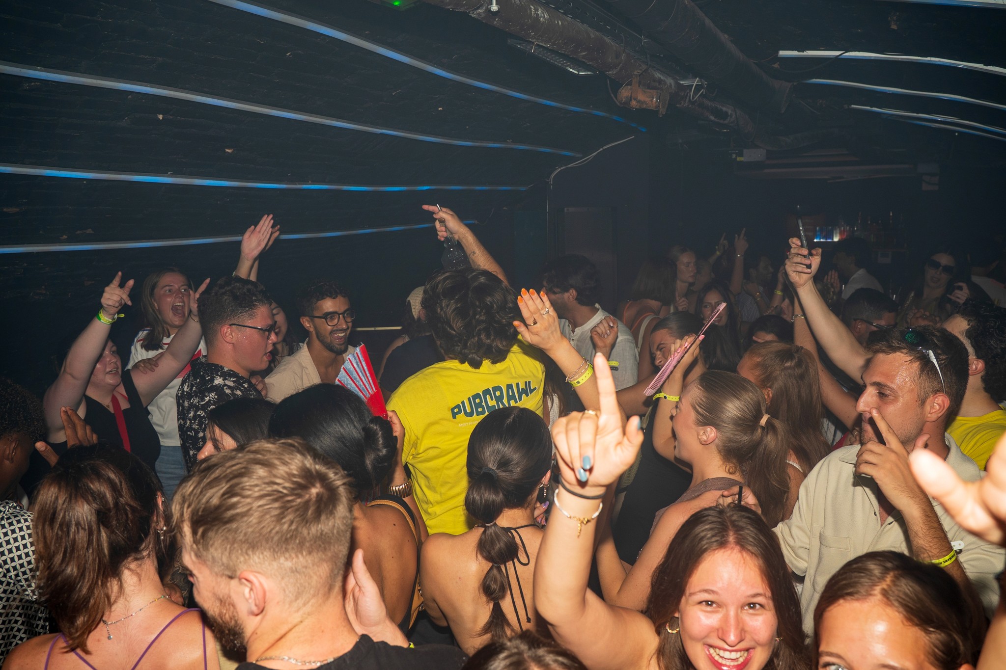 A lively bar in Nice at night with people singing along, hands in the air, colorful lights and a DJ booth in the background.
