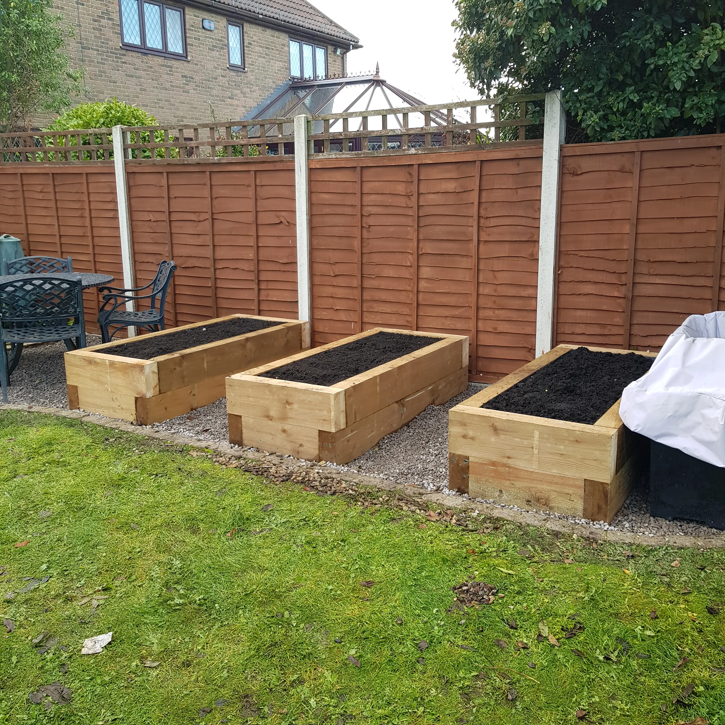 Three wooden raised garden beds are arranged in a backyard, surrounded by grass and a wooden fence.