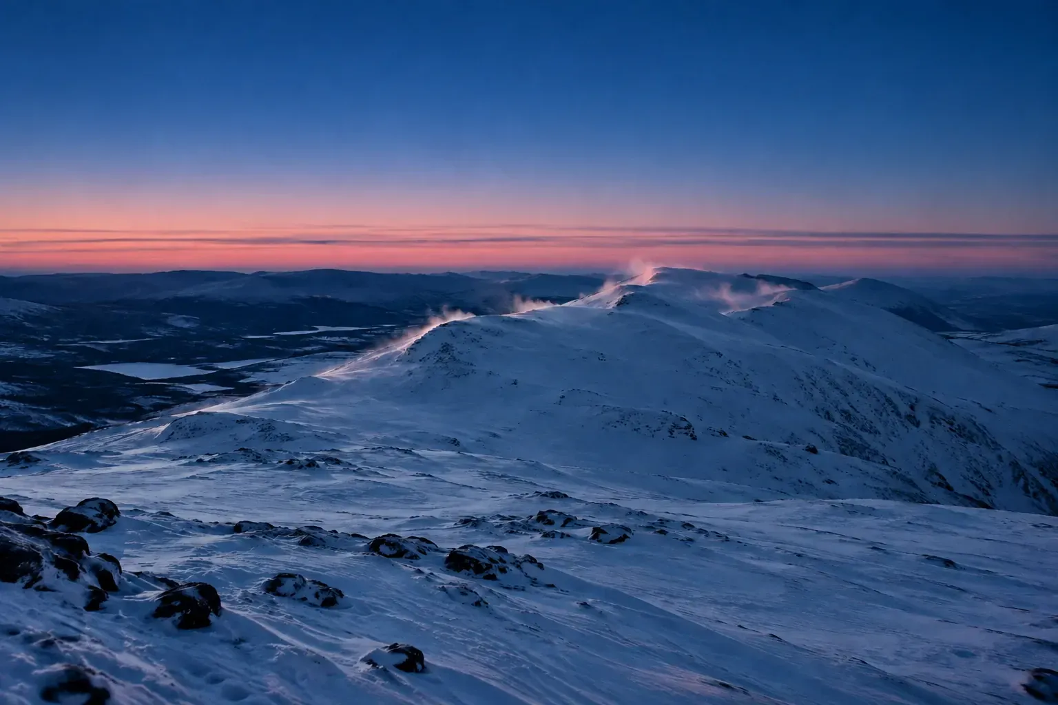 A long snow-covered fell ridge with wind-blown snow, a thin pale-pink band along the horizon under a deep blue sky.