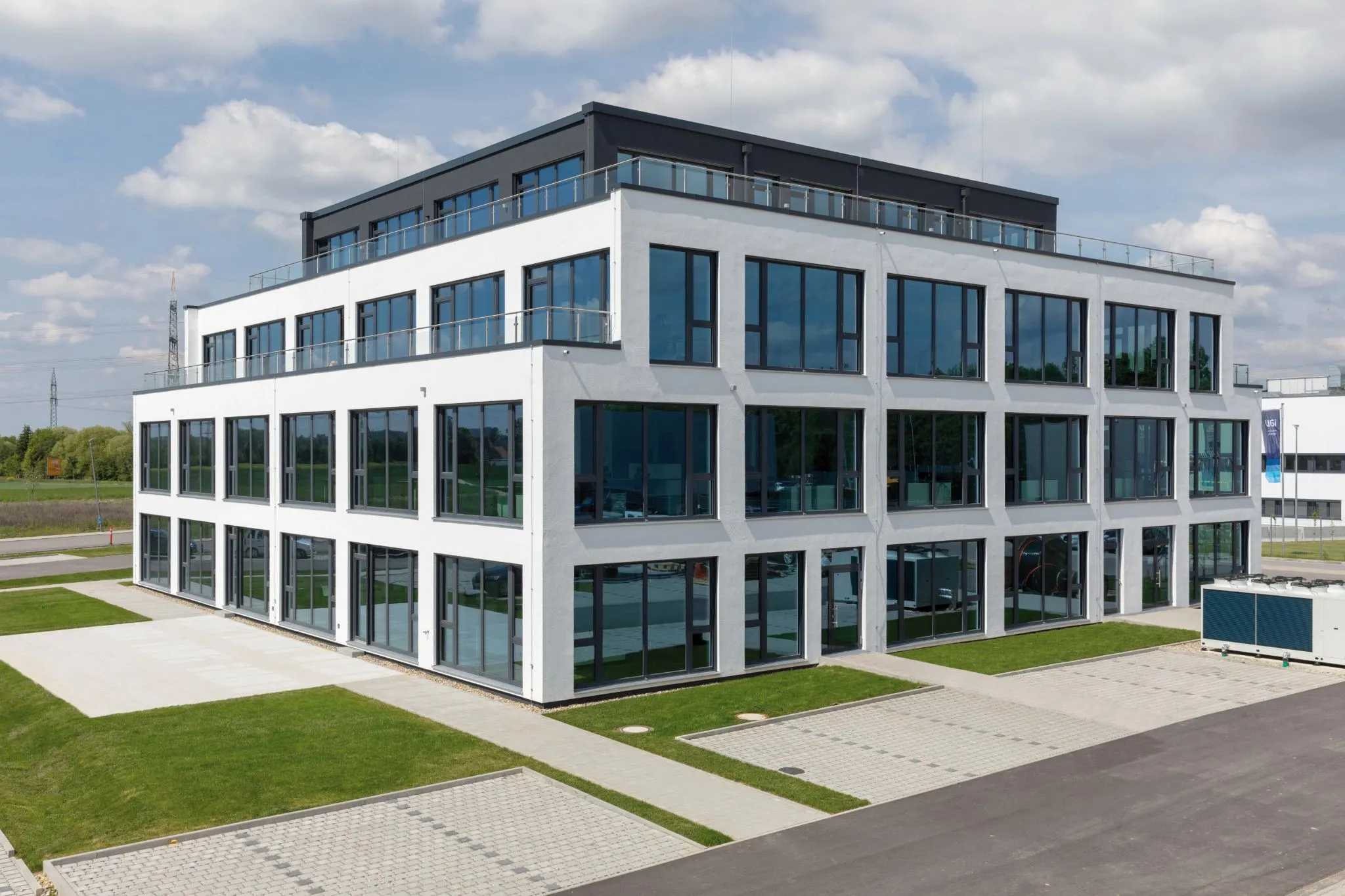 Weißes Bürogebäude mit dunklem Dachaufbau. Große Fensterfronten und Glasbalkone auf mehreren Etagen im Sommer.
