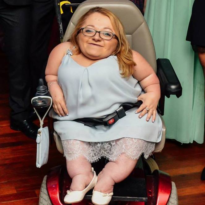 Gem in wheelchair smiling in a pale blue dress with a matching back hanging from controller