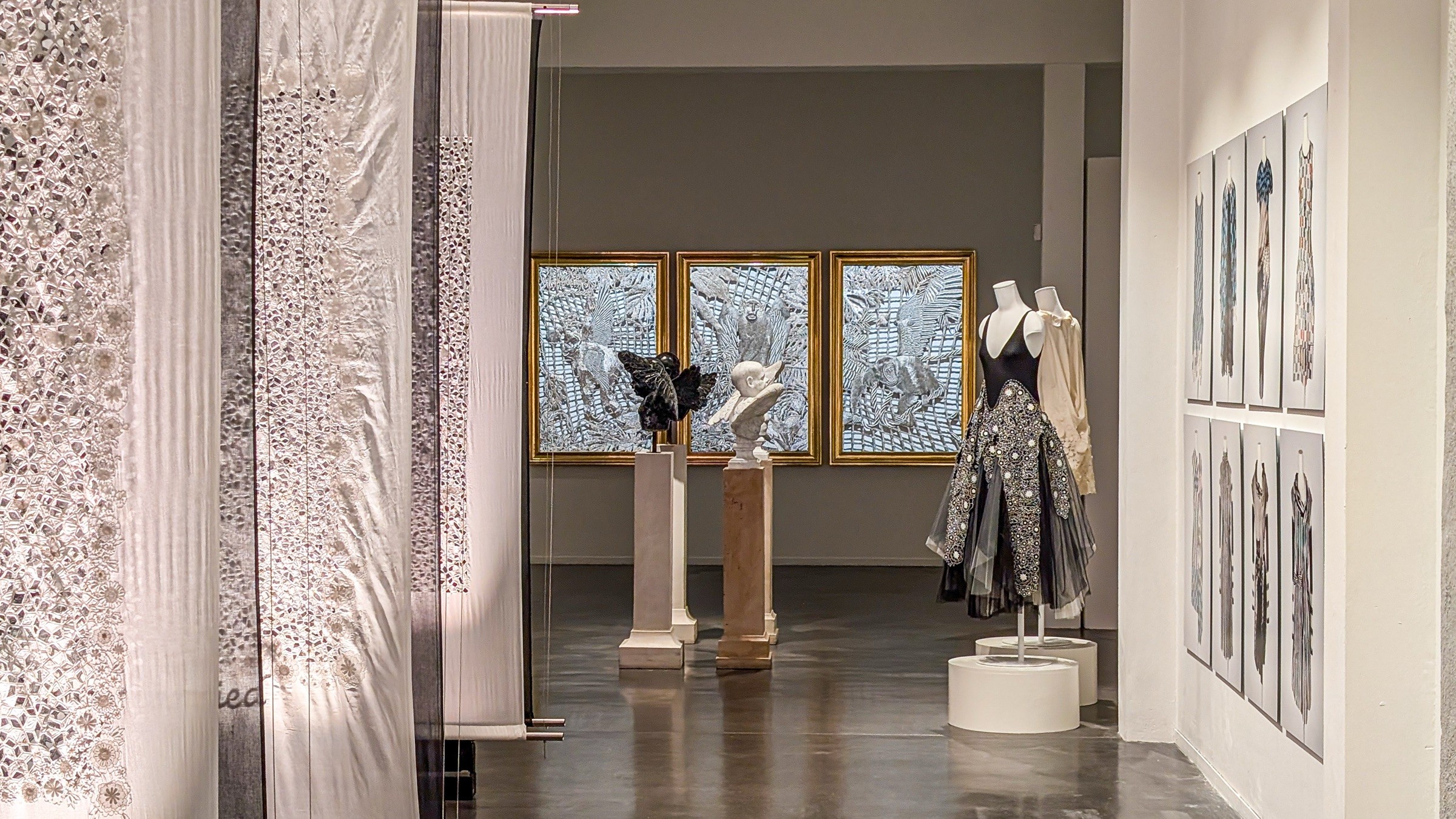 An exhibition room with a mannequin in a black dress, framed drawings on the wall and a curtain of light