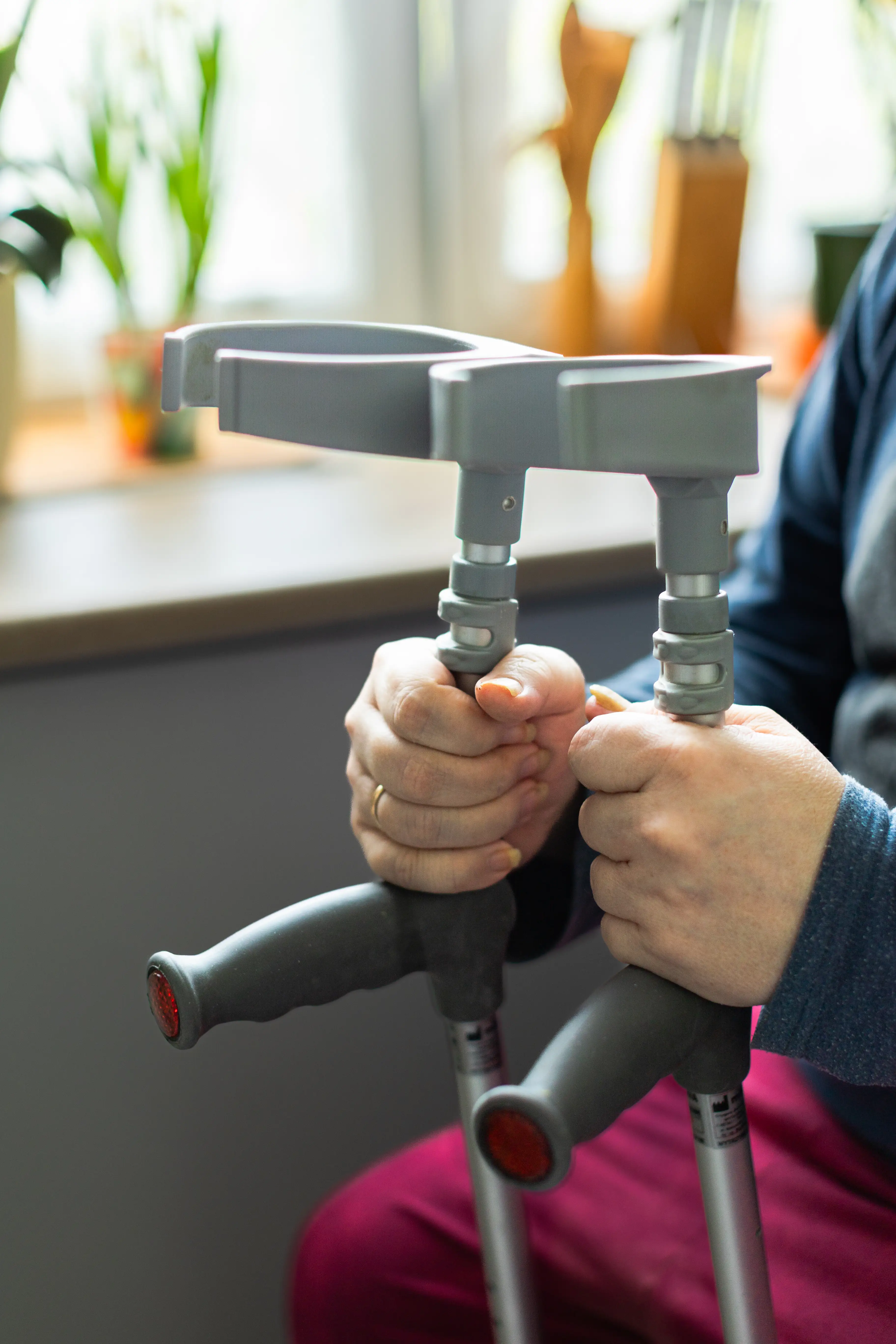 Dłonie zaciśnięte na kulach dla niepełnosprawnych. W tle kolana osoby podpierającej się na kulach oraz okno z parapetem.