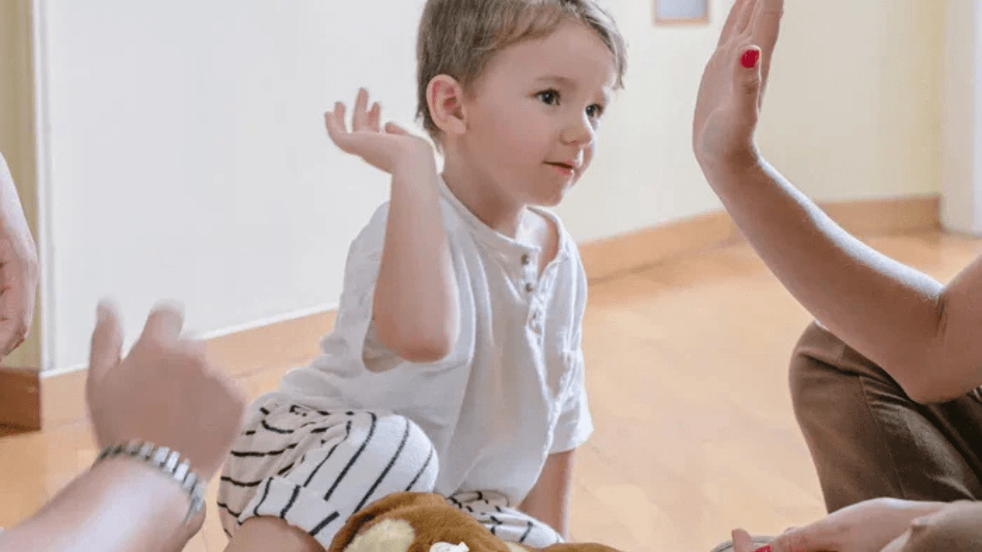 Young child sitting on the floor raising a hand for a high-five during a playful interaction with adults