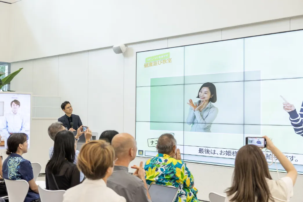 Japanese municipal officials taking photos of an ESTsoft AI Human demonstration for senior care services on a large display