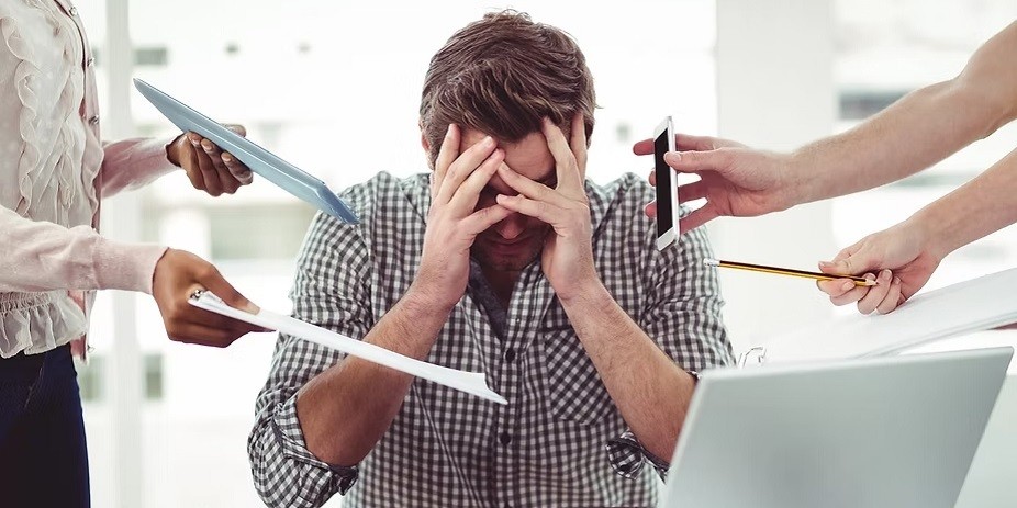 An overwhelmed businessman sitting at a desk with his head in his hands, surrounded by multiple arms holding tablets, phones, and documents representing extreme workplace stress and team inefficiency.