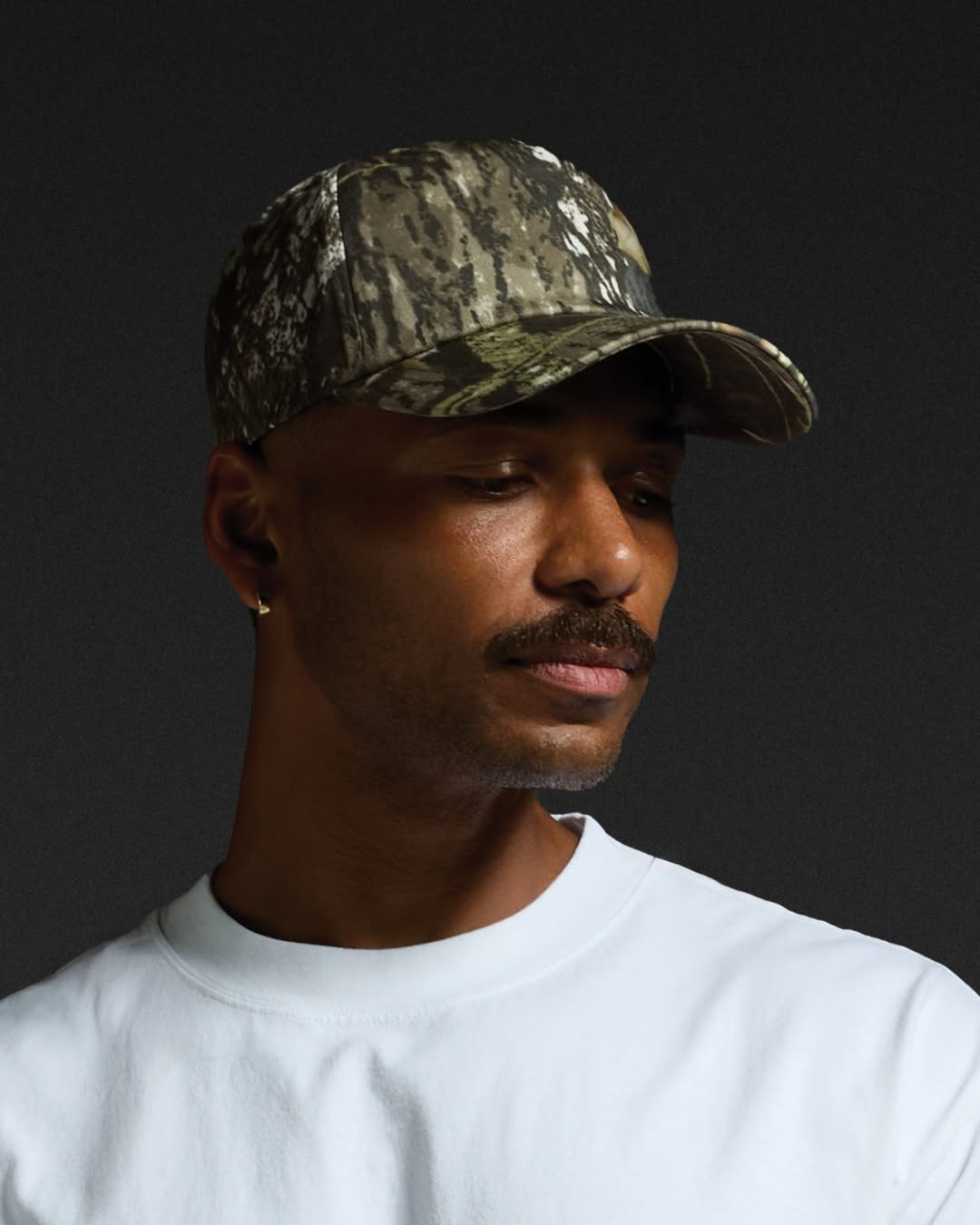 A man in a camo hat looking to the right wearing a white shirt against an almost black background.