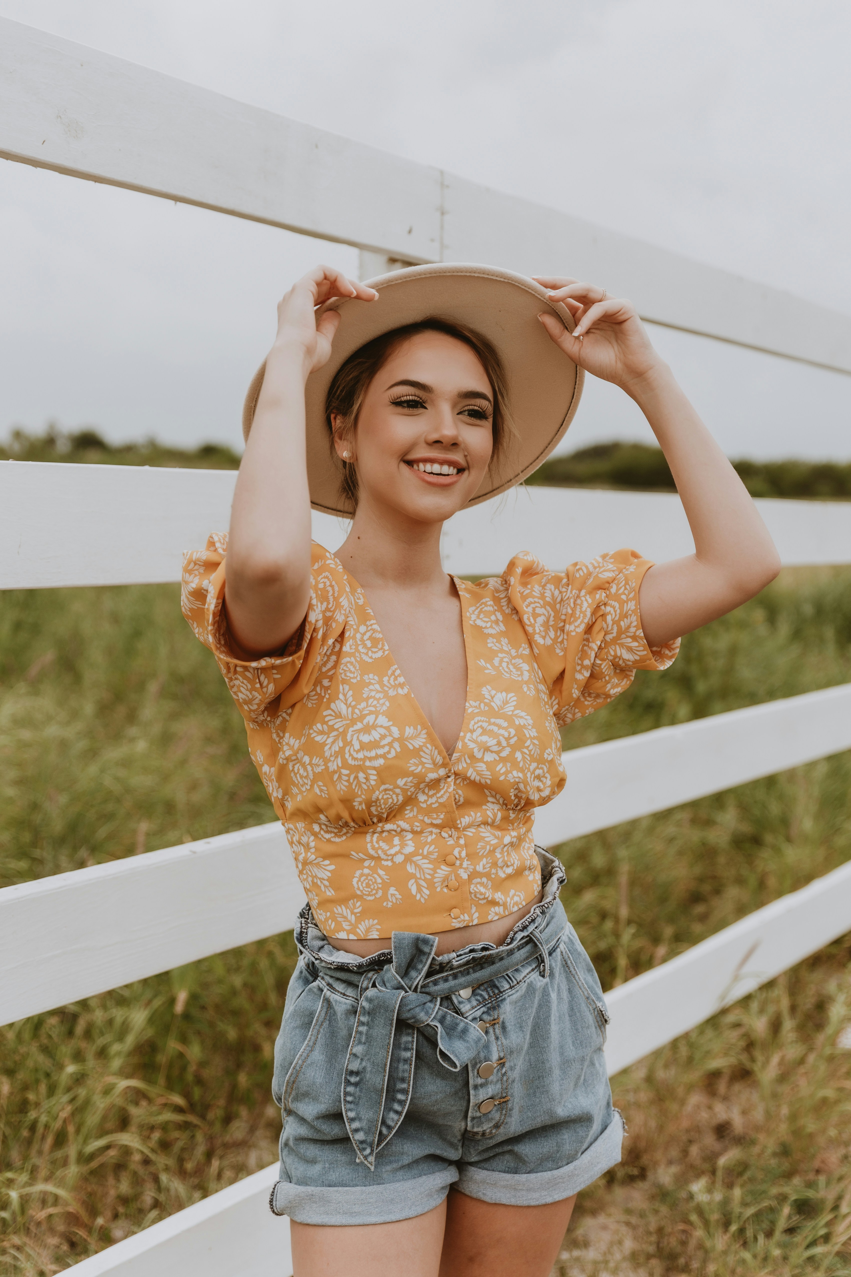 A woman in a yellow top and denim shorts stands by a white fence, smiling and enjoying the outdoors.