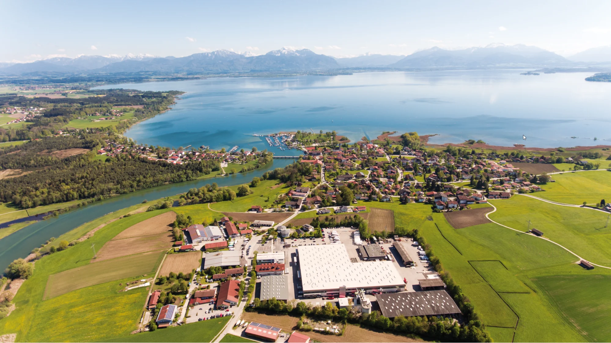 Luftaufnahme eines Sees mit Bergkulisse, einem ruhigen Fluss, einer Siedlung am Ufer und Gewerbebauten auf grünen Feldern.