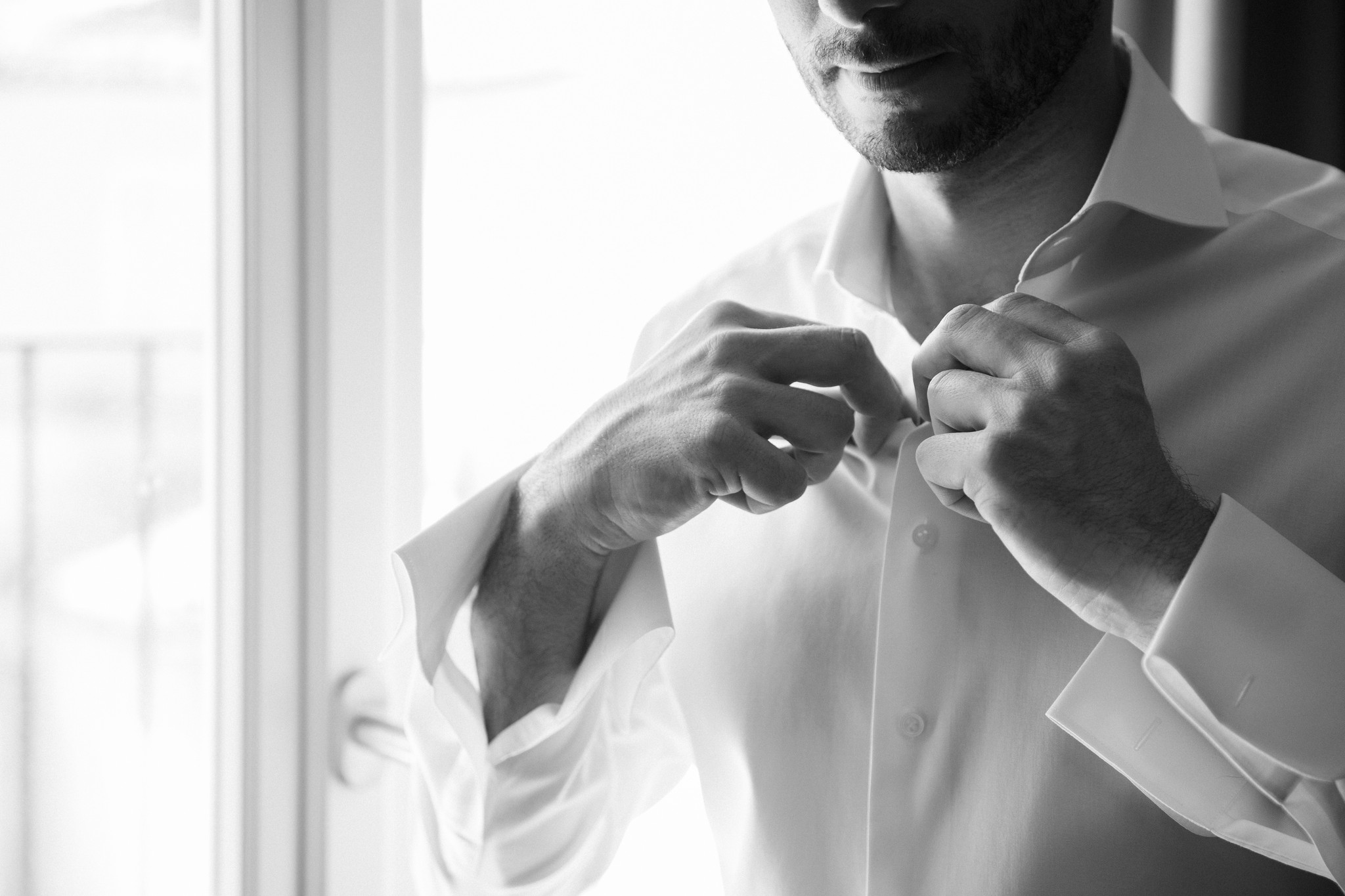 Sposo allaccia i bottoni della camicia bianca in bianco e nero, preparativi di matrimonio con luce naturale dalla finestra