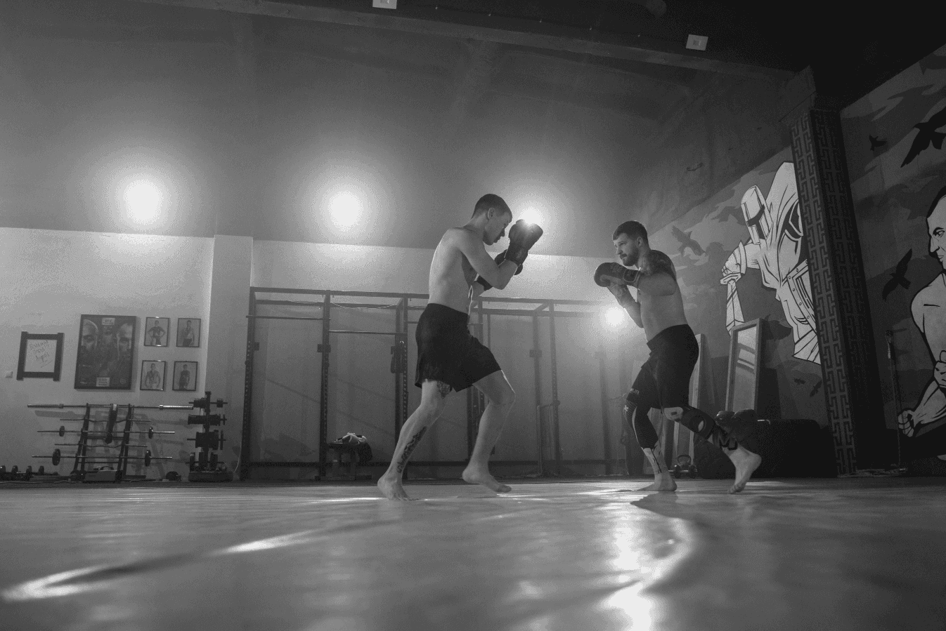 Two barefoot fighters sparring during MMA or kickboxing training inside a gym, facing each other on padded mats under bright lights.