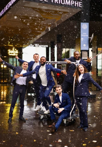 A group of cheerful people in formal attire poses energetically in front of an upscale hotel entrance, adorned with decorative signage, as gentle snowfall creates a festive and lively atmosphere.