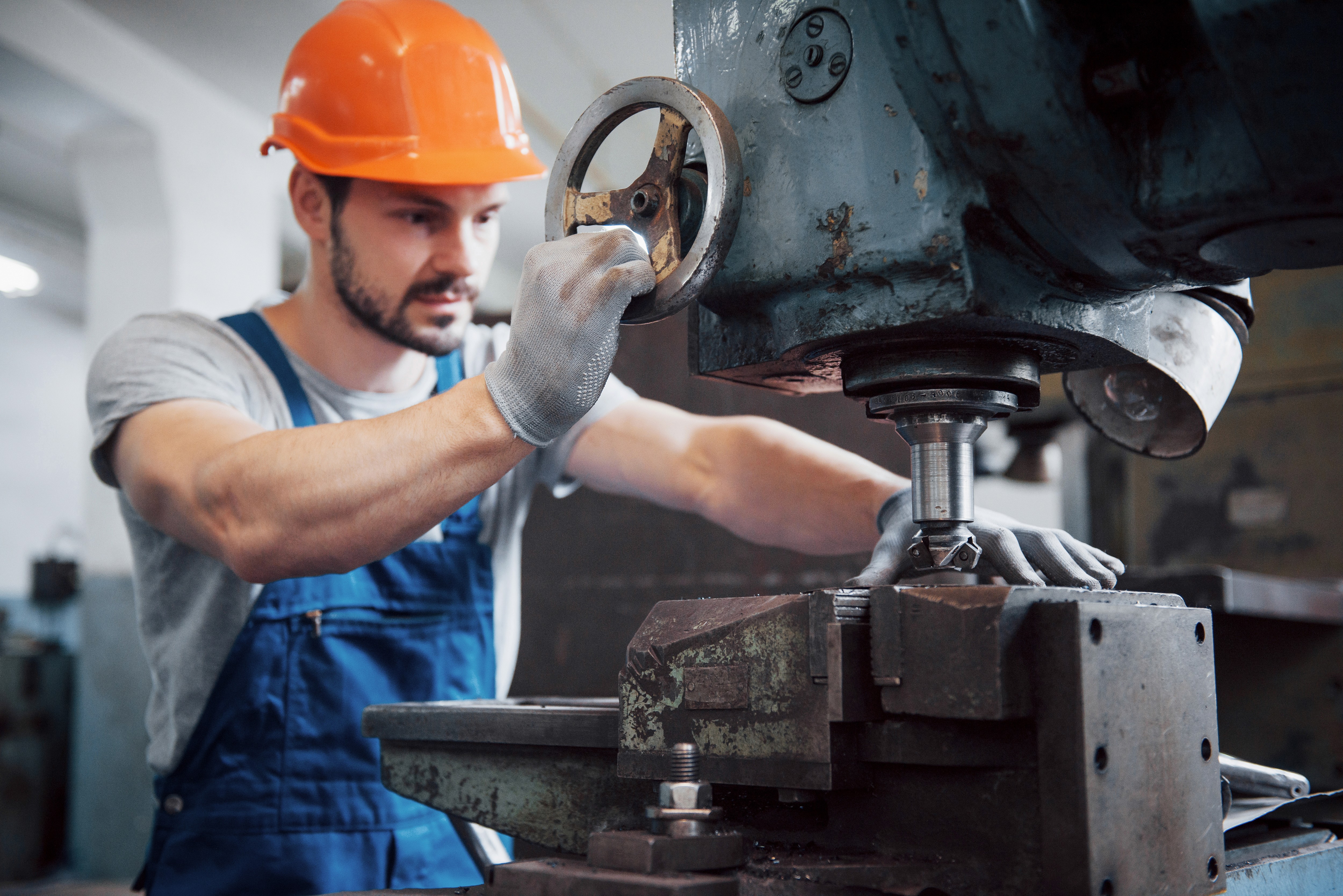 Man in a hard hat and blue overalls operates a metalworking machine in an industrial workshop. He appears focused and skillful, adjusting a wheel.