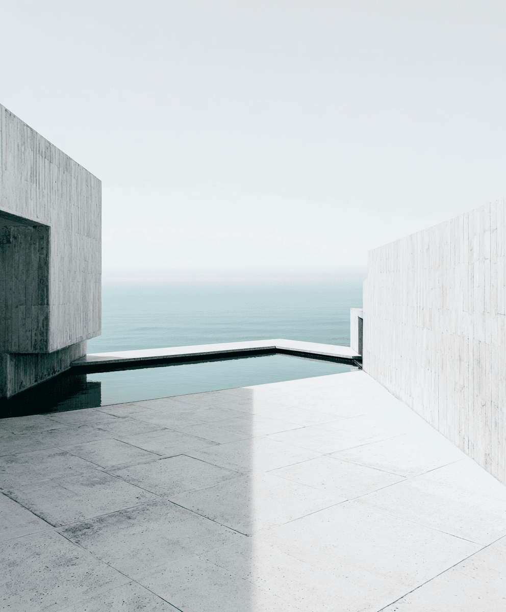 A pale concrete terrace with sharp geometric lines frames a still, angular pool that opens onto a calm, mist-hazed sea under a soft white sky.