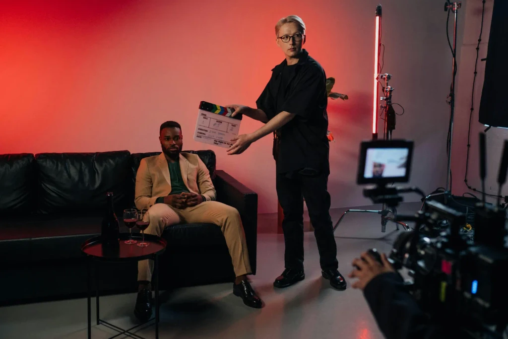 In a red-lit studio, a crew member holds a clapperboard before a man in a tan suit on a couch, seen behind a camera.