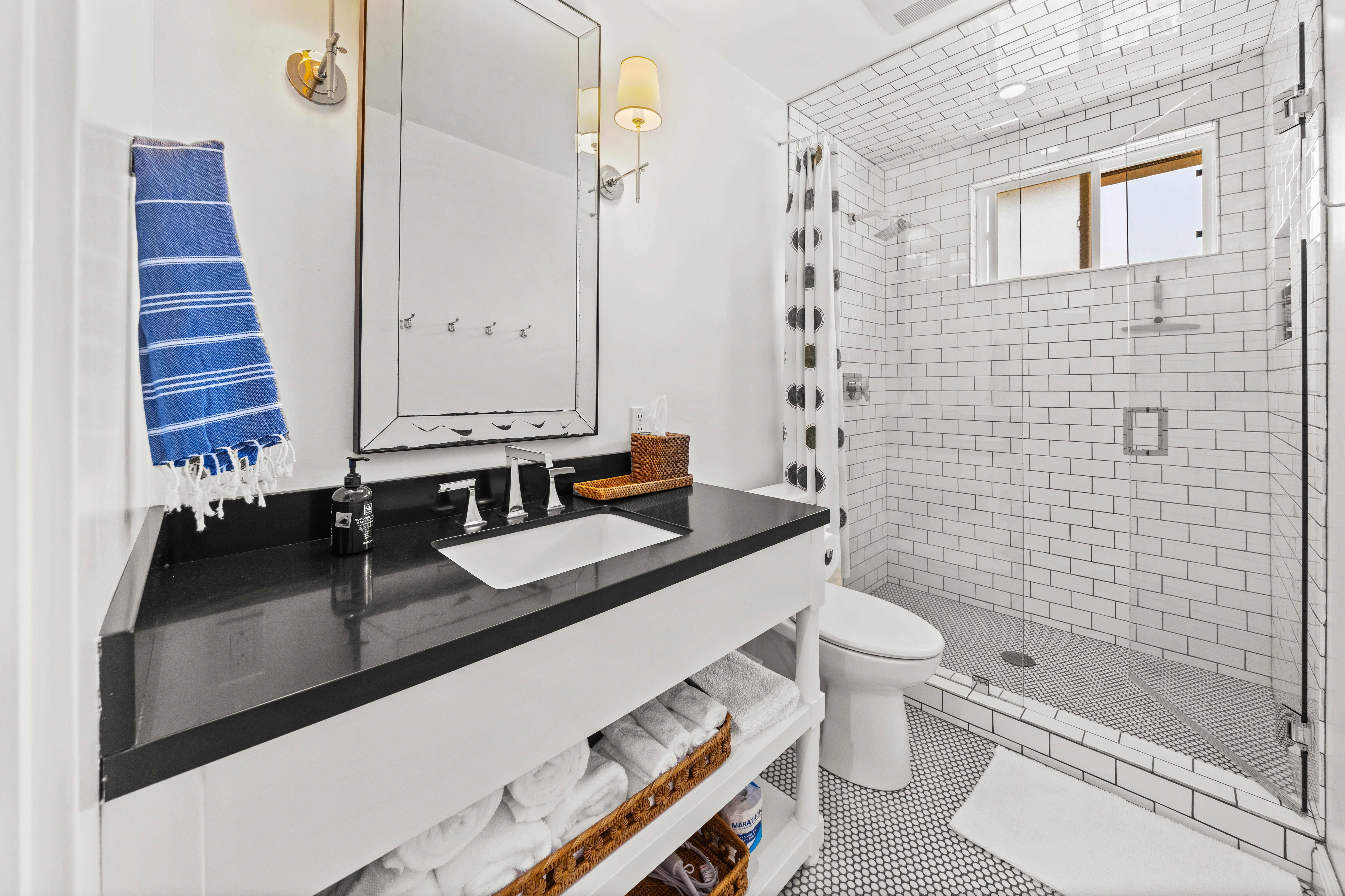 A bright, modern bathroom featuring a sleek black countertop, open shelving, and a spacious subway-tiled shower.