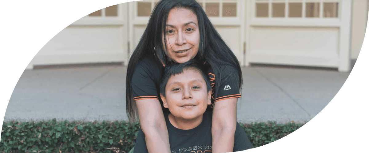 Uma mulher sorridente de cabelos longos e escuros, vestindo uma blusa preta, abraça seu filho pequeno, que também sorri, enquanto posam juntos do lado de fora de um prédio com janelas de moldura branca.