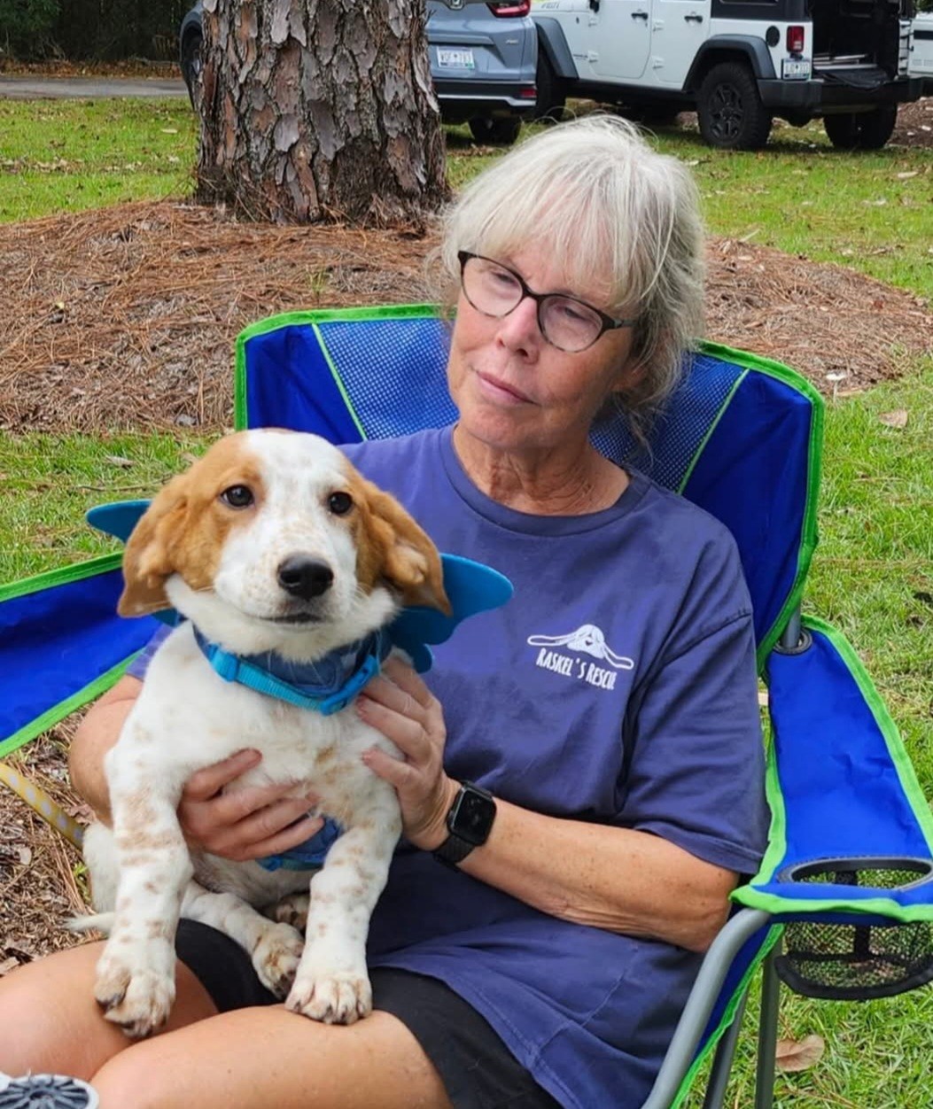 An older woman in a blue chair holds a puppy in her lap, both looking at the camera in a green outdoor setting.