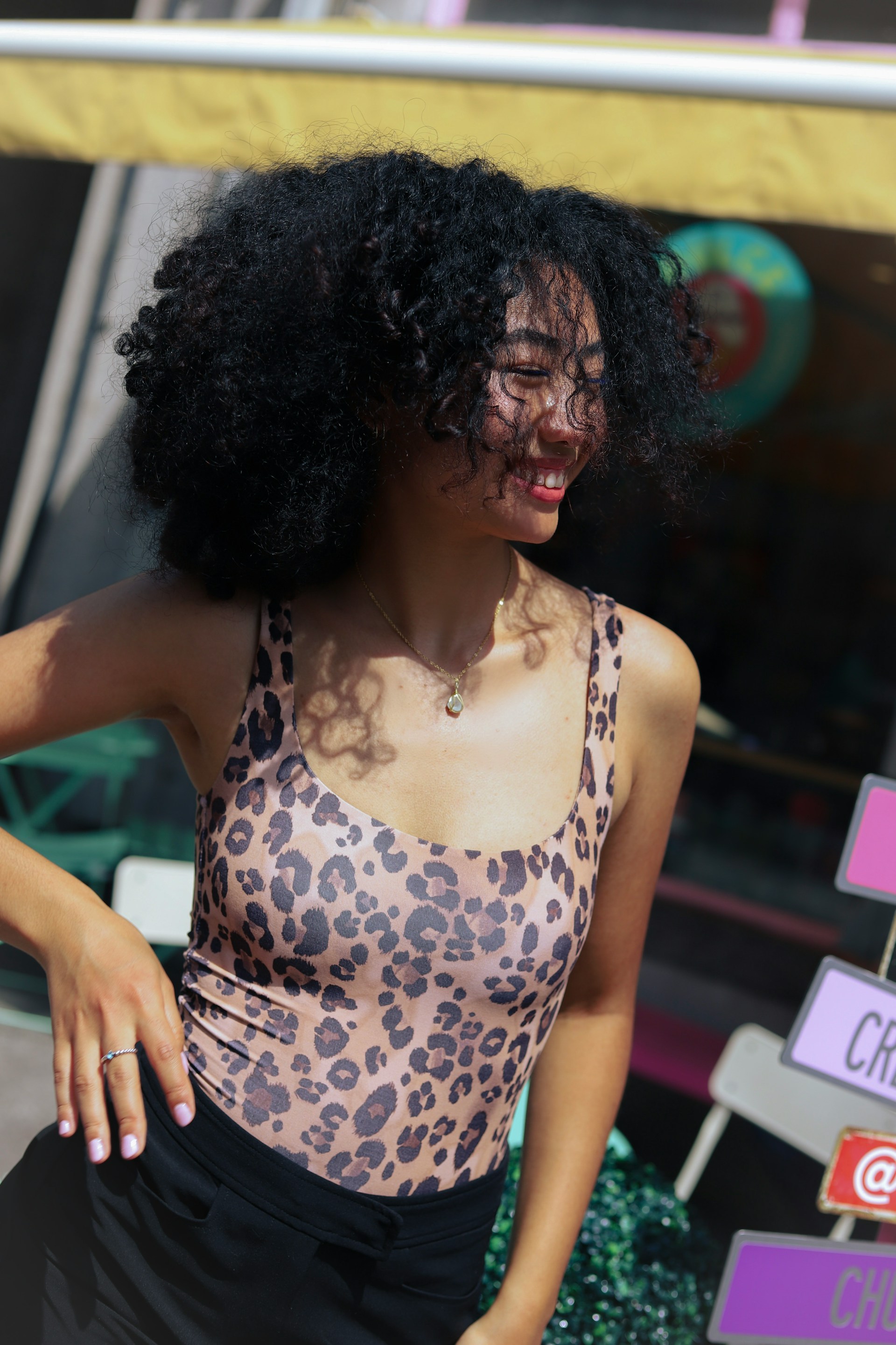 A candid shot of a woman with voluminous, curly black hair laughing and looking away. She is wearing a leopard-print tank top and a gold pendant necklace in a sunlit outdoor setting.