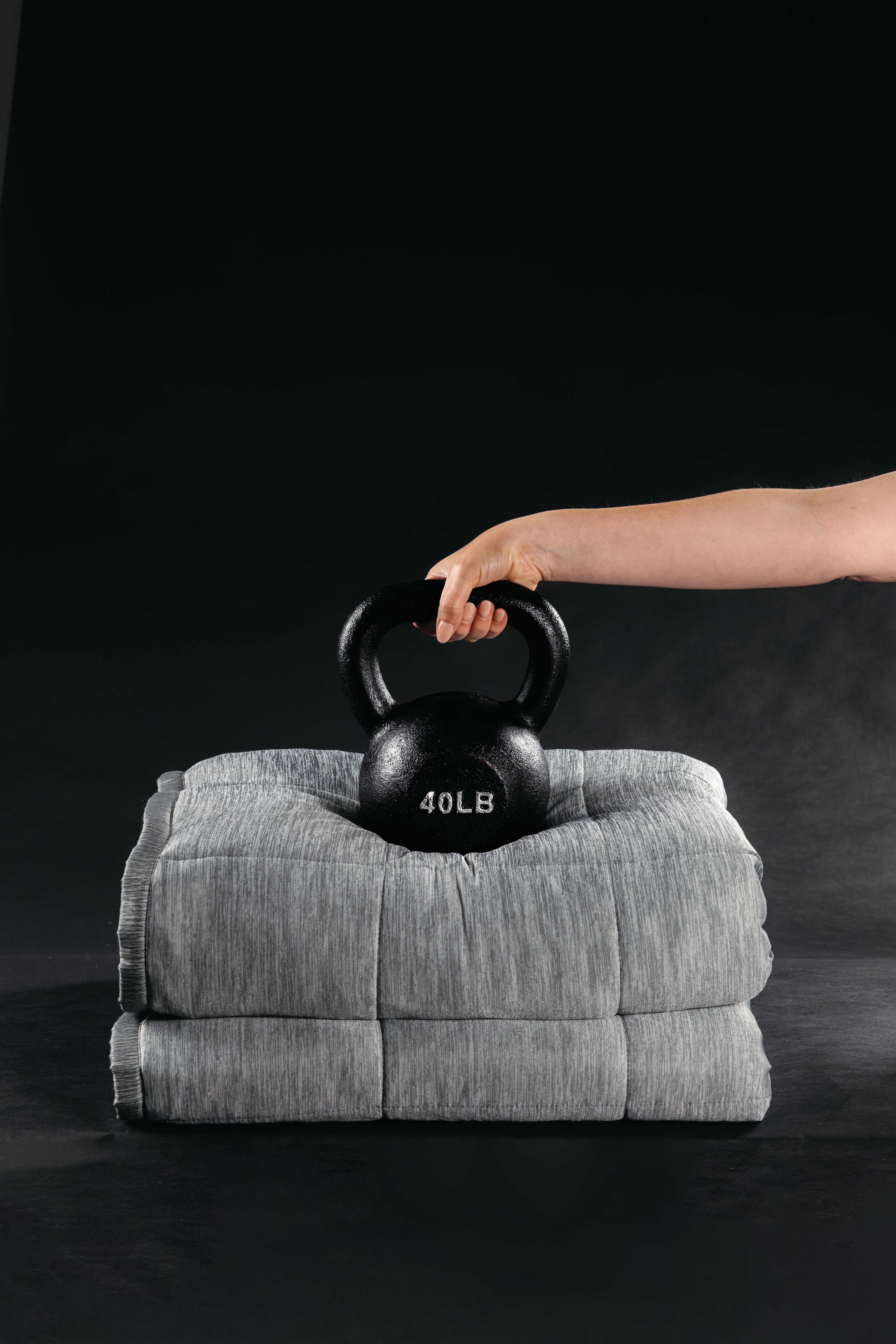 A hand setting a 40 lb kettlebell on a folded grey XL Weighted Big Blanket against a black backdrop