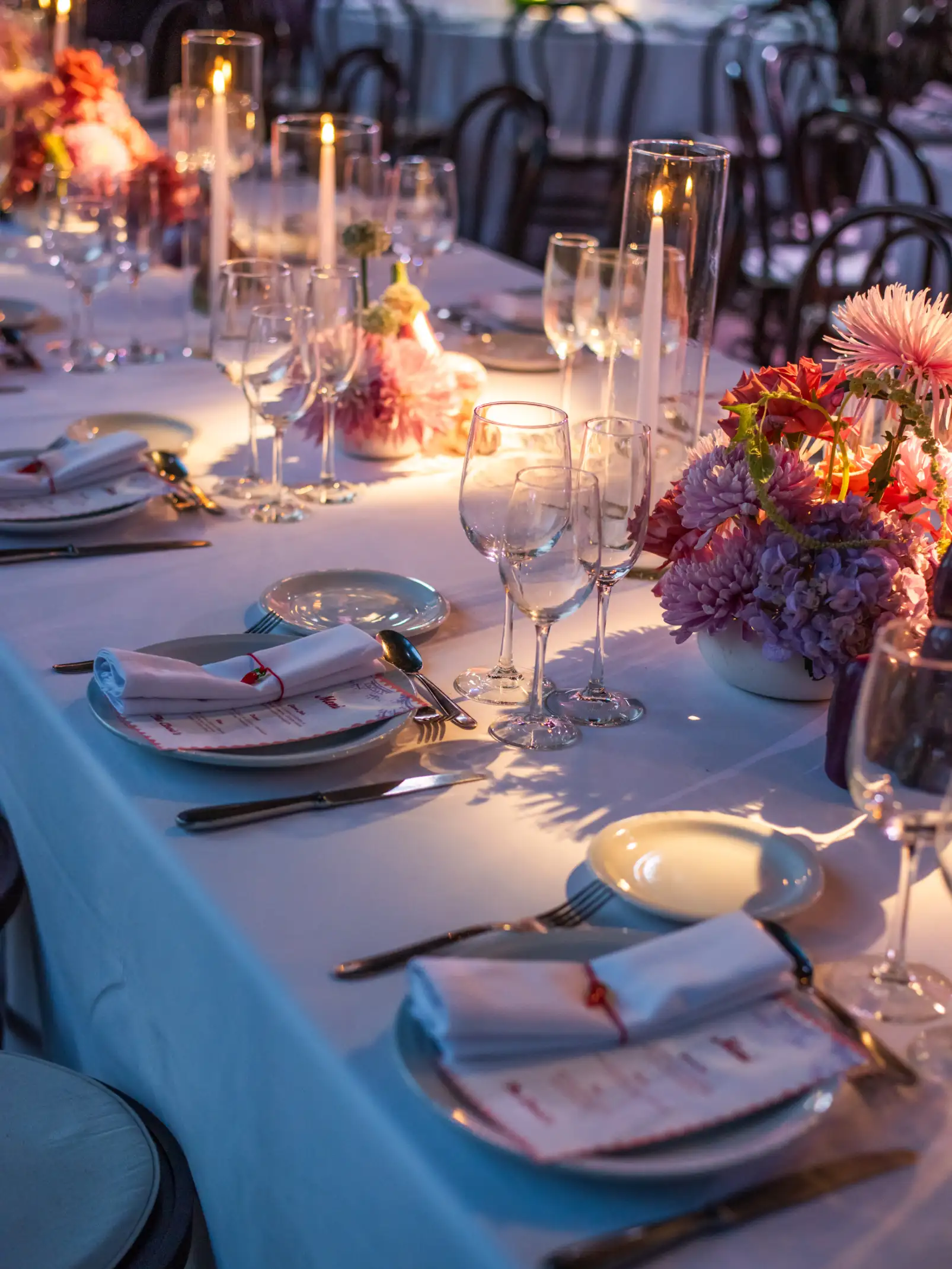 Mesa larga con centros florales en tonos pastel, velas altas y vajilla blanca bajo luz cálida de evento nocturno.