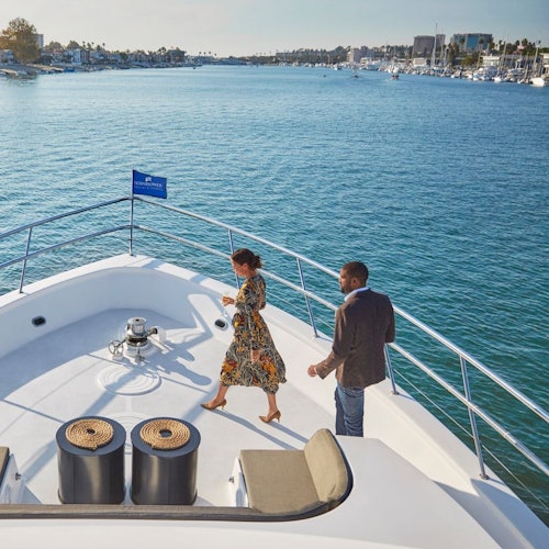 A woman and man walk on the deck of a yacht on a sunny day, with a marina and boats in the background.