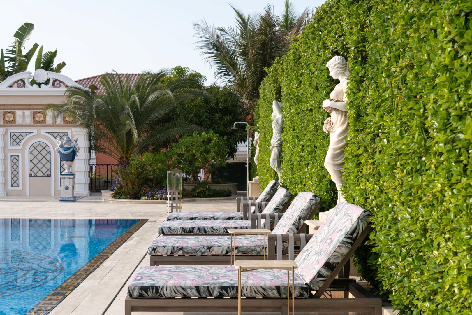 Poolside loungers with floral cushions line a luxurious swimming pool, surrounded by manicured hedges, statues, and palm trees, creating a serene resort atmosphere.