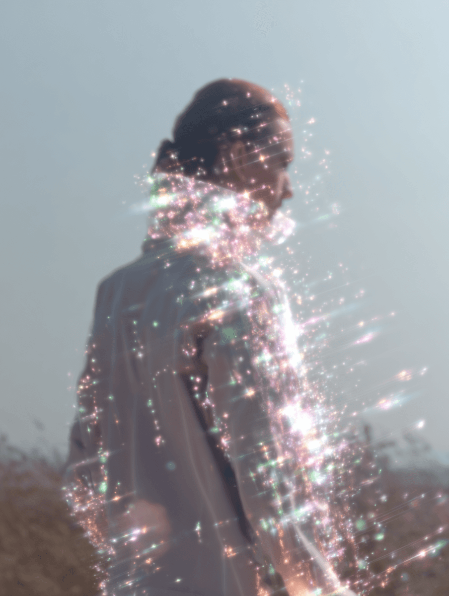 A person stands with their back turned, illuminated by soft sunlight and surrounded by shimmering reflections.