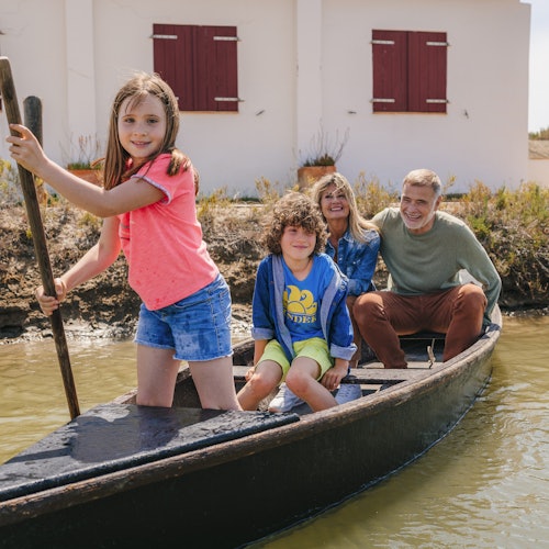Eine Familie mit vier Personen, zwei Erwachsene und zwei Kinder, genießt eine Bootsfahrt auf einem schmalen Fluss in der Nähe eines weißen Gebäudes mit roten Fensterläden.