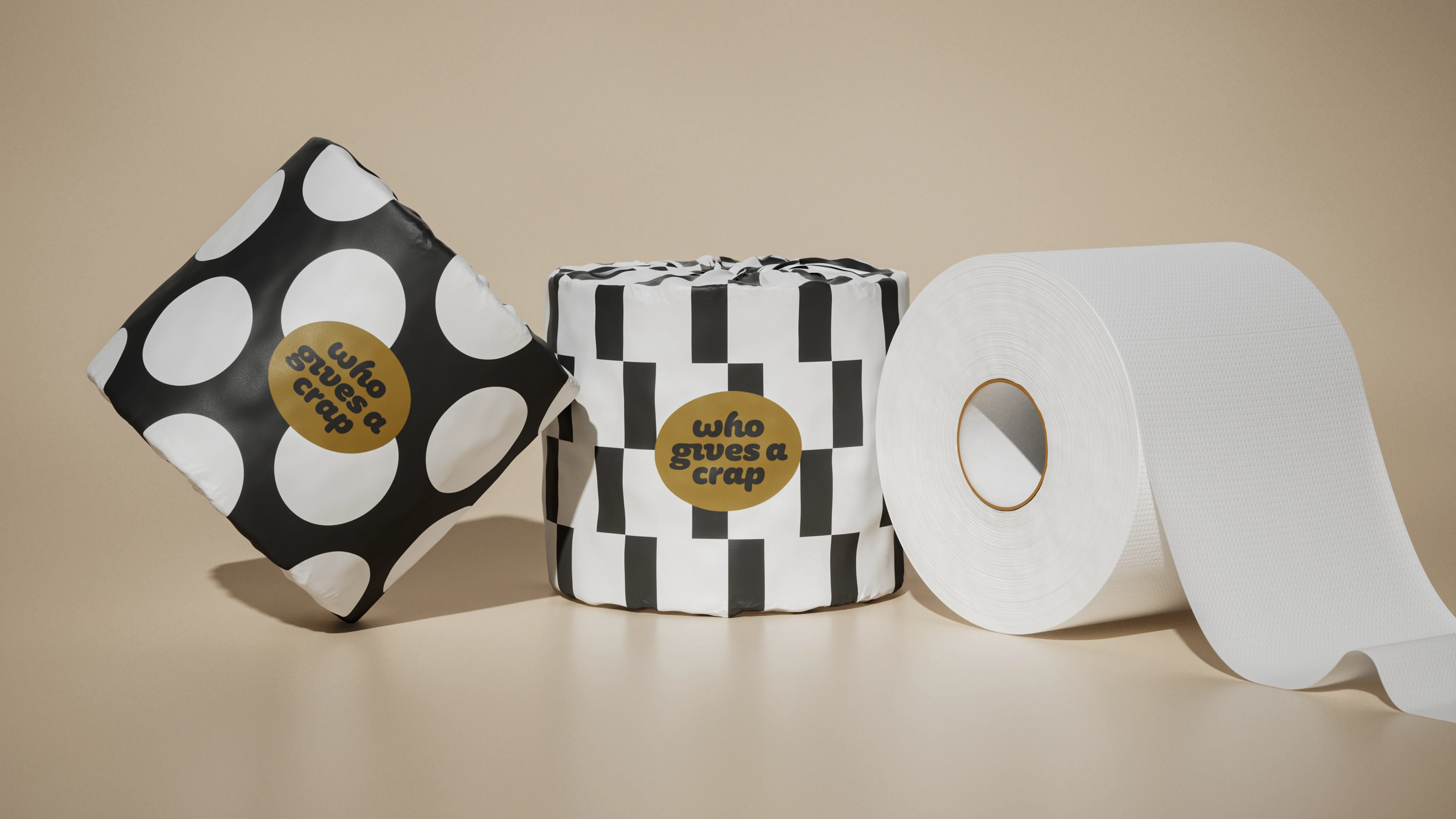 Three Who Gives A Crap rolls on a beige backdrop: two wrapped in black-and-white dot and check patterns, one unwrapped white roll