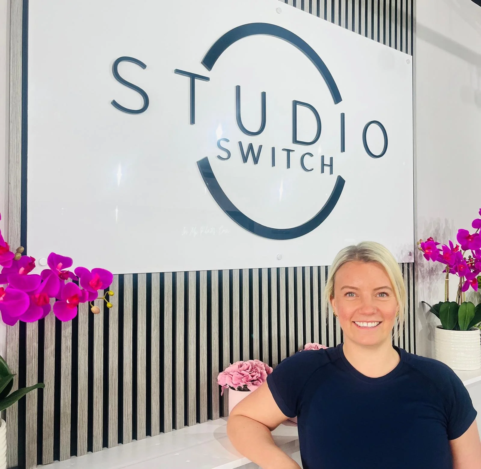 Laura, a Studio Switch Pilates instructor, smiling in front of the Studio Switch reception sign and pink orchids.