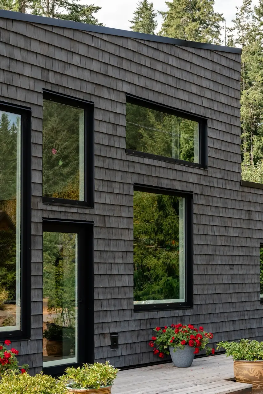 Modern two-story home with large glass windows, warmly lit interiors, and a balcony. Surrounded by trees and a manicured lawn at dusk. Cozy ambiance.